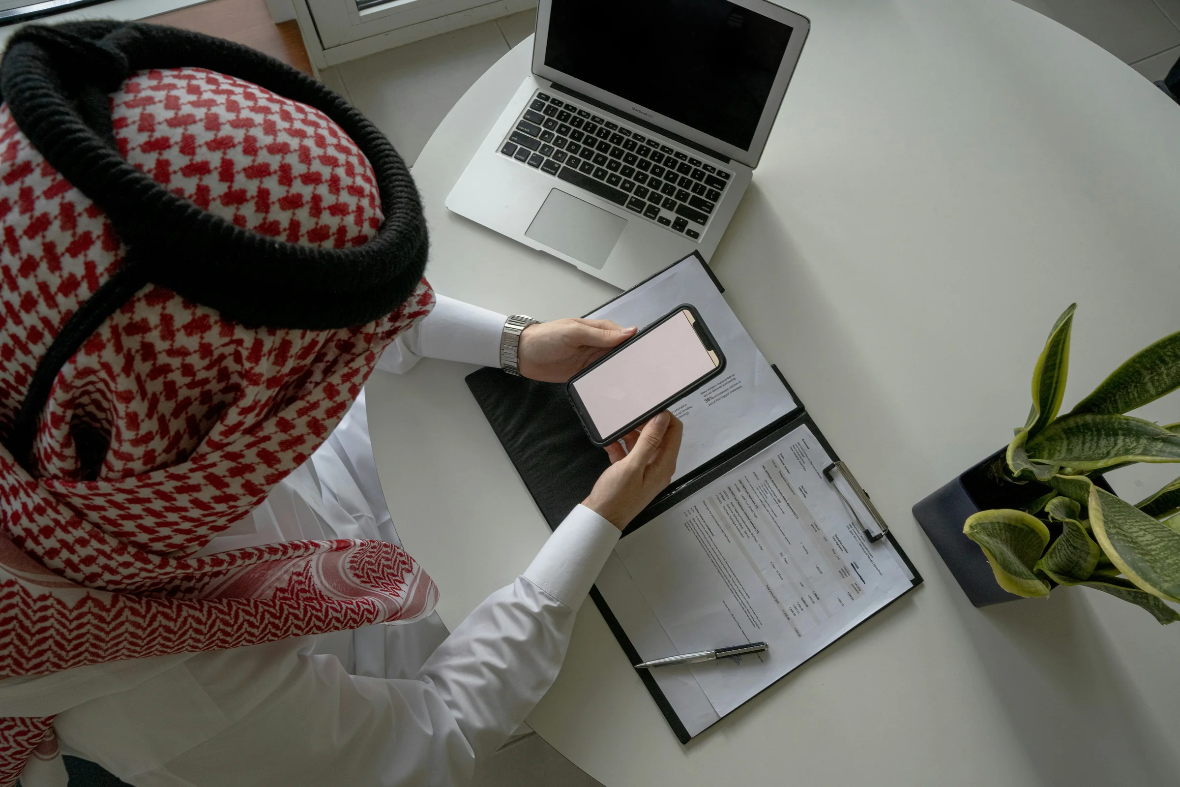 A man in a ghutra holding his phone over a folder of forms